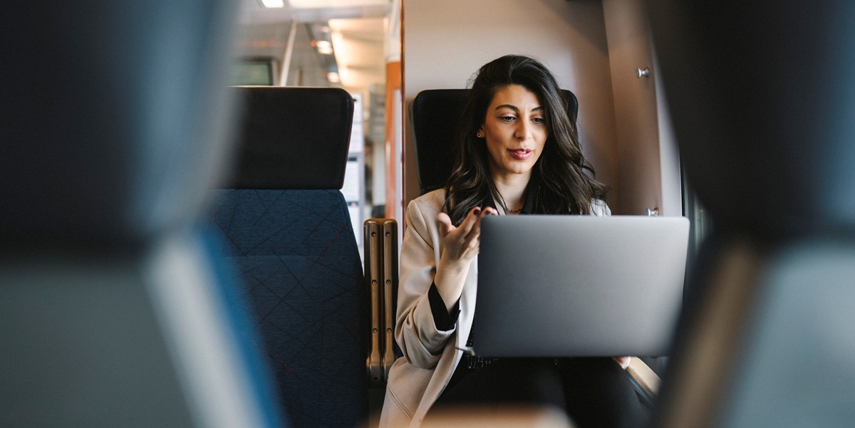 a finance leader is seated in transit on a train with a laptop on her lap; she appears to be speaking while in a meeting or call.