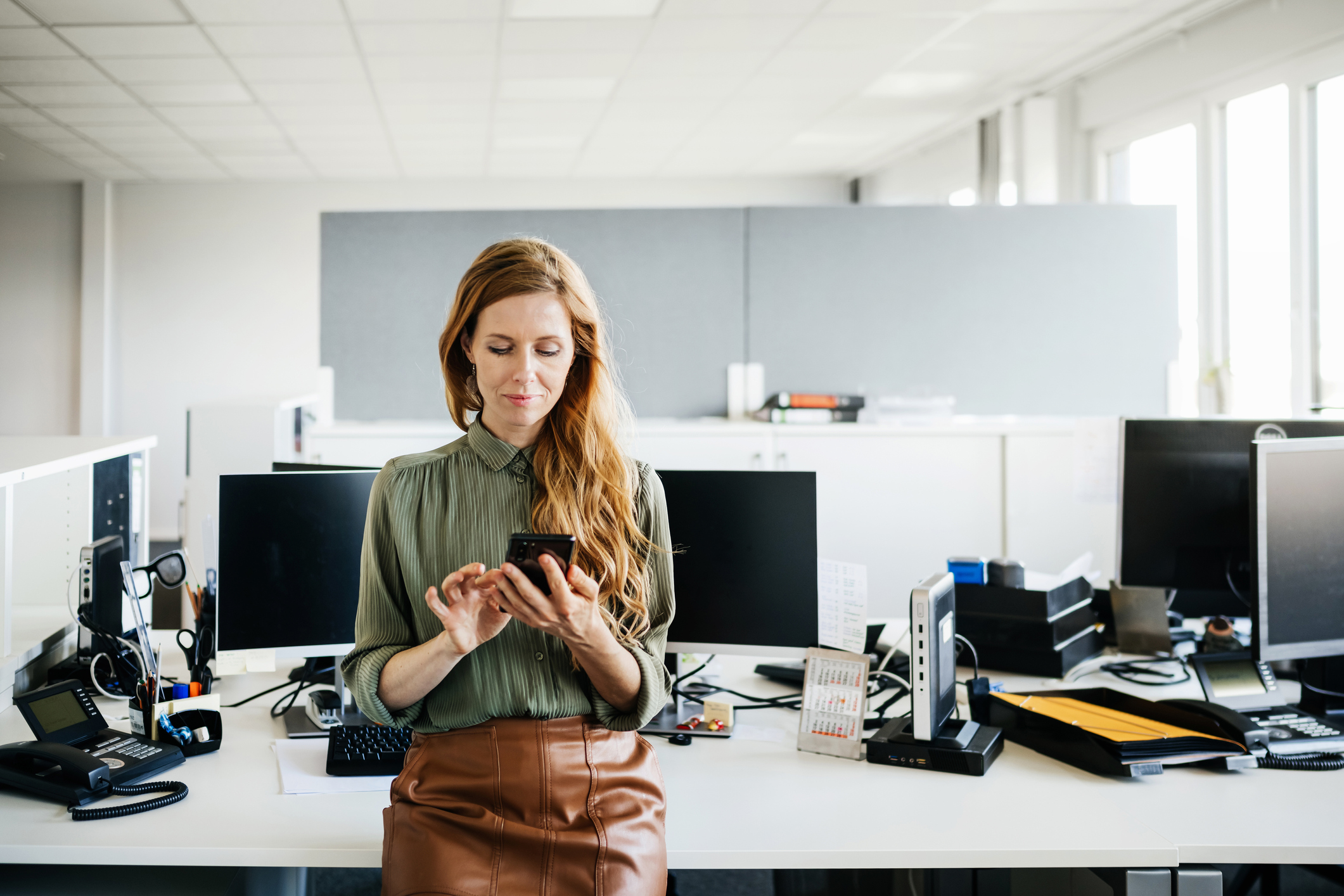 Woman leaning against a desk of computers looking at her phone
