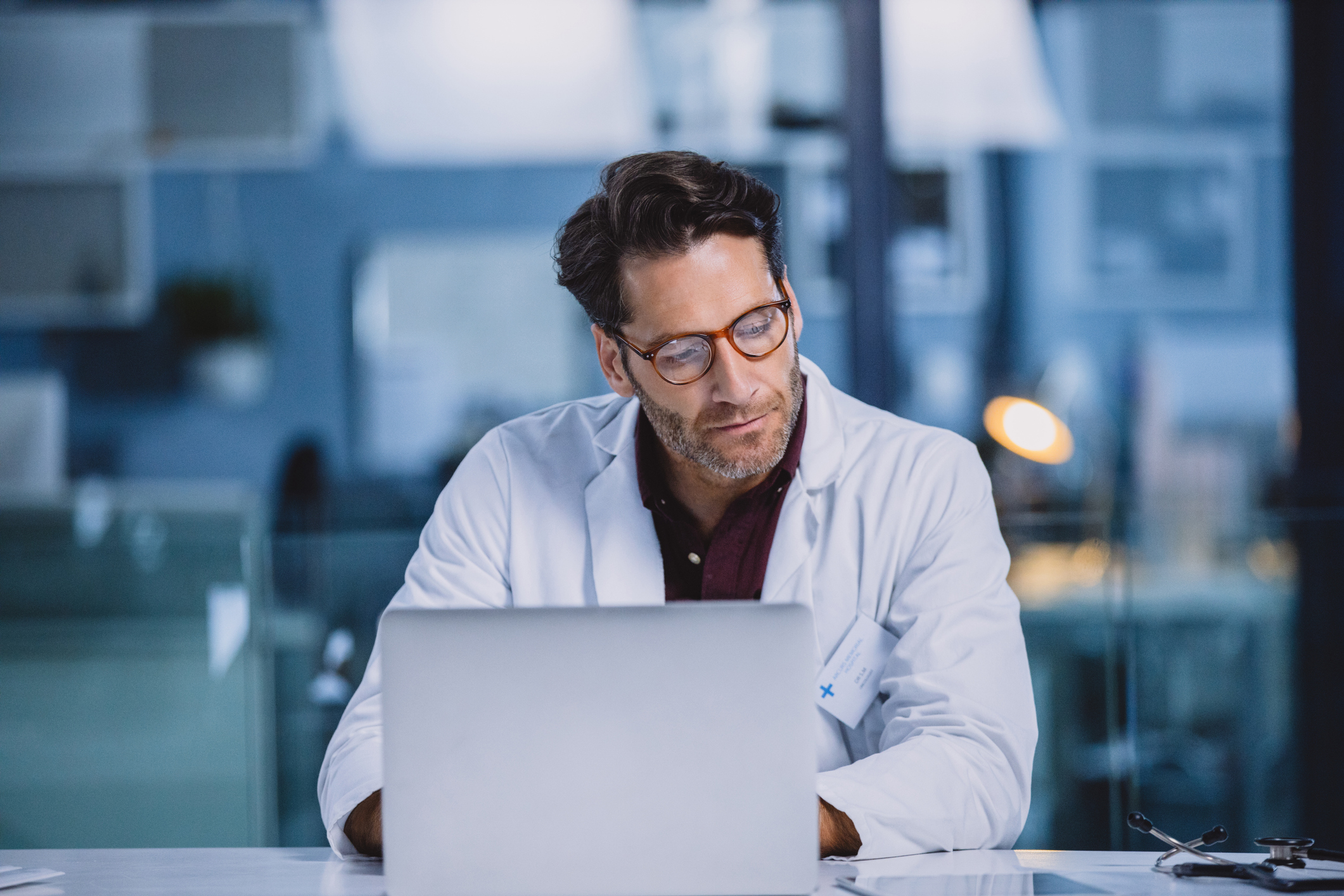 Male doctor sitting looking at paperwork with a laptop open
