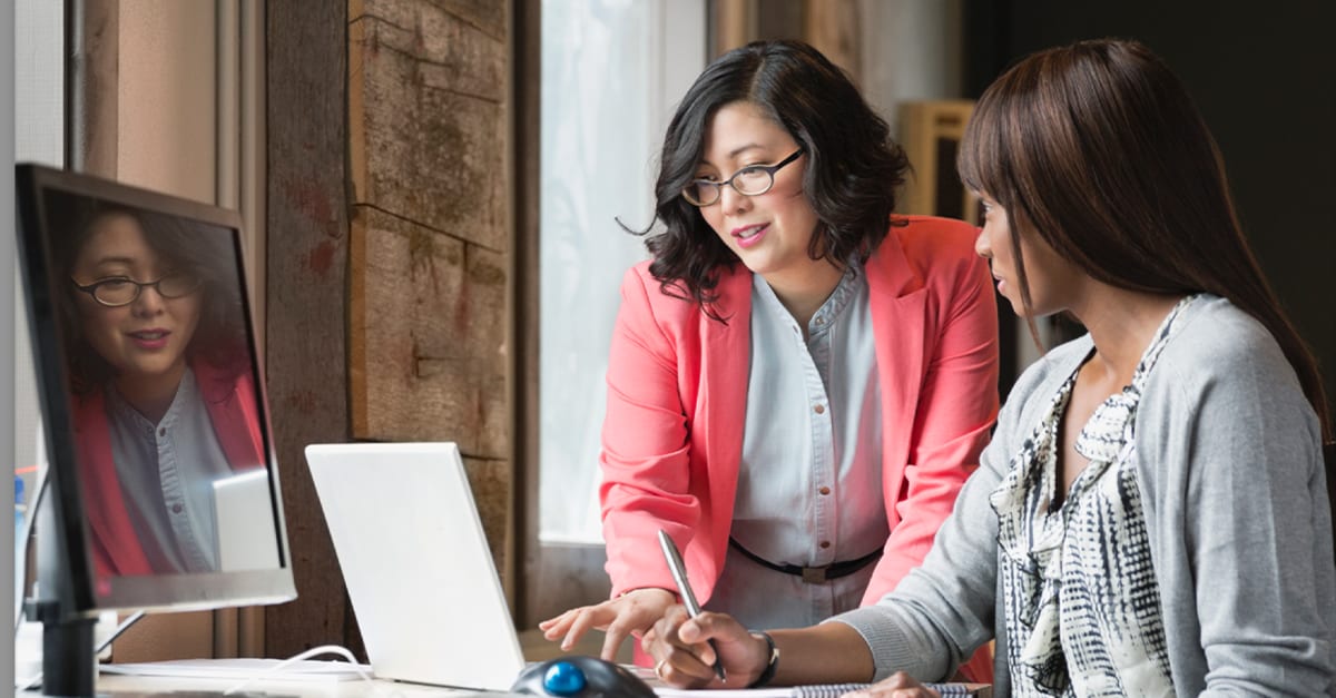 Woman seated at an office computer desk collaborates with another woman standing next to her